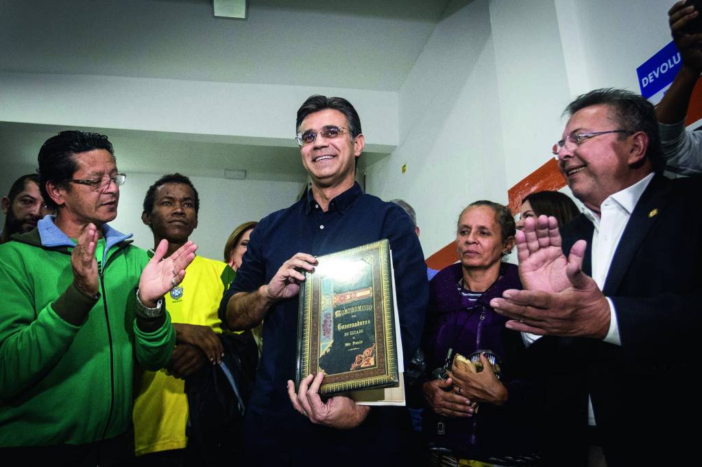 Imagem mostra homem sorrindo em volta de pessoas que aplaudem. Ele sorri.