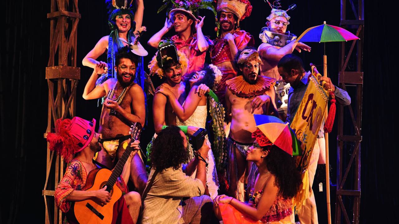 Grupo de atores com roupas coloridas, alguns com chapéus de frevo, contracenando no palco