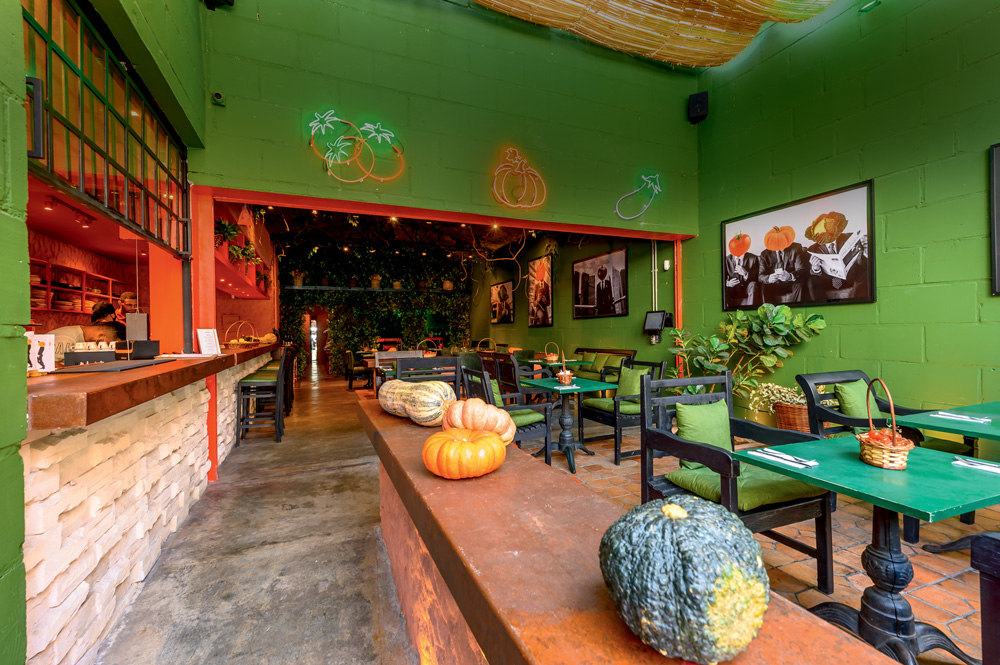 Ambiente de restaurante decorado por mesas de madeira pintadas de verde, cadeiras de cor escura, parede de cor verde e mais