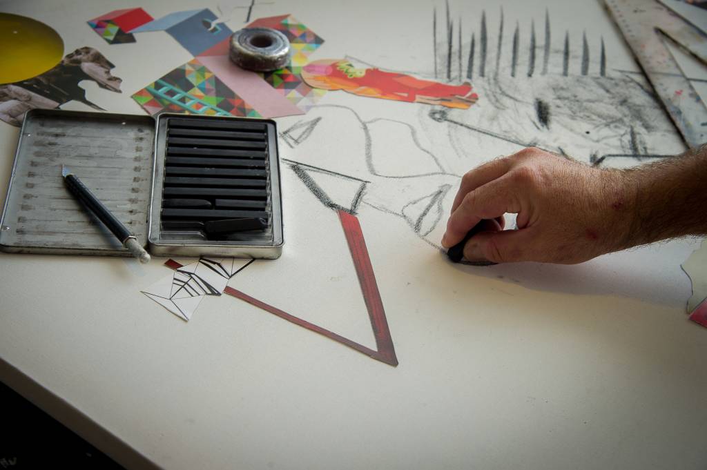 Imagem exibe detalhe de tela branca com alguns desenhos, lápis e a mão de Vímerson trabalhando na obra.