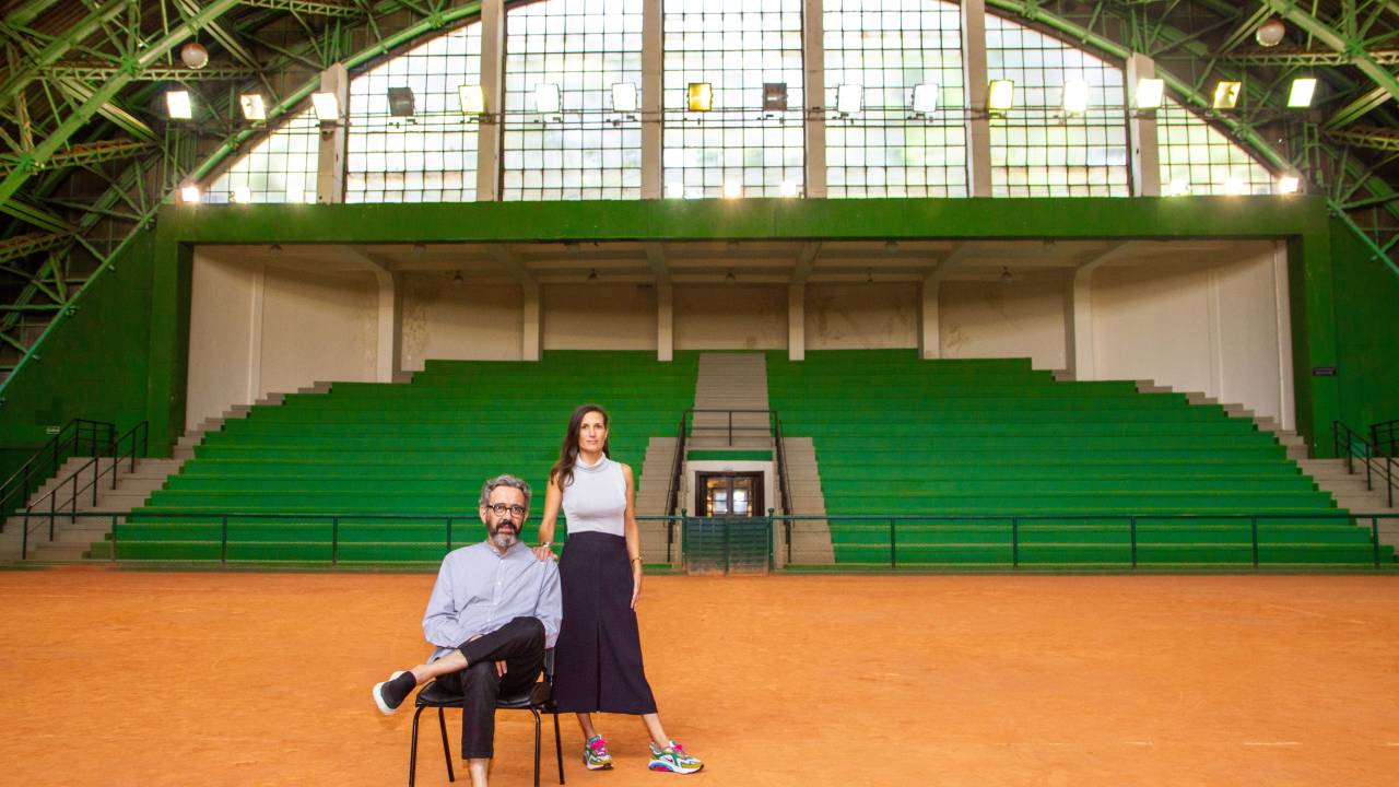 Waldick Jatobá e Camilla Barella, das feiras Made e ArPa, posam em um grande estádio com chão cor de areia. Ele está sentado de pernas cruzadas e ela de pé ao lado, com saia preta e blusa sem mangas branca.