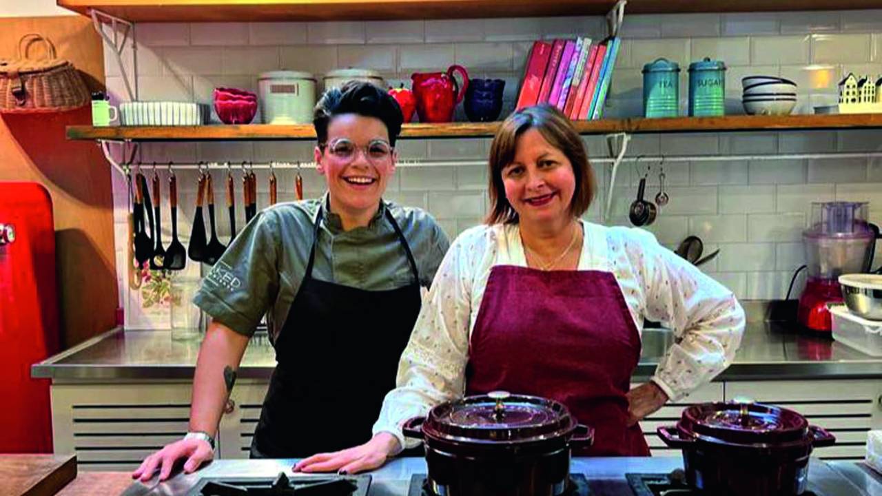 Cristiane Skrings e Carla Pernambuco posam lado a lado usando aventais de diferentes cores em um ambiente de cozinha