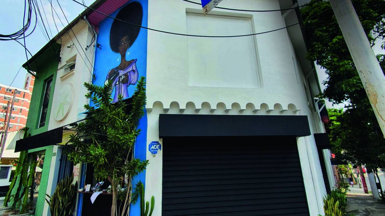 Fachada de cafeteria instalada em uma esquina com parede de cor branca, portão de cor preta e pintura na lateral esquerda sobre fundo de cor azul