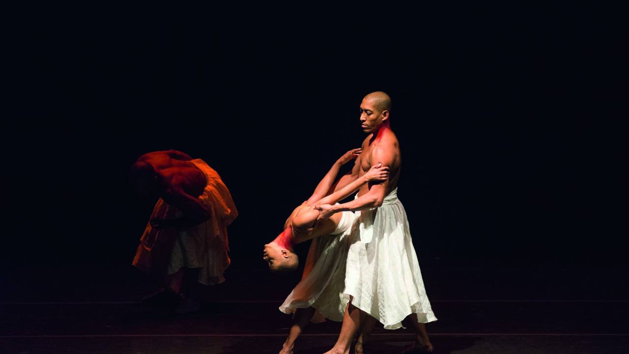 Imagem mostra duas pessoas em palco vestindo saias brancas, dançando. Uma terceira pode ser vista ao fundo, no escuro.