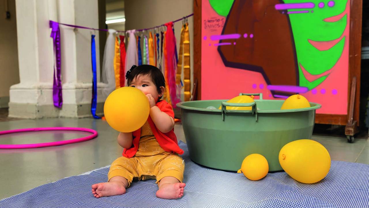 Um bebê está sentado no chão de uma sala e brinca com um balão amarelo, que coloca na boca. No fundo, uma bacia verde tem balões amarelos flutuando na água