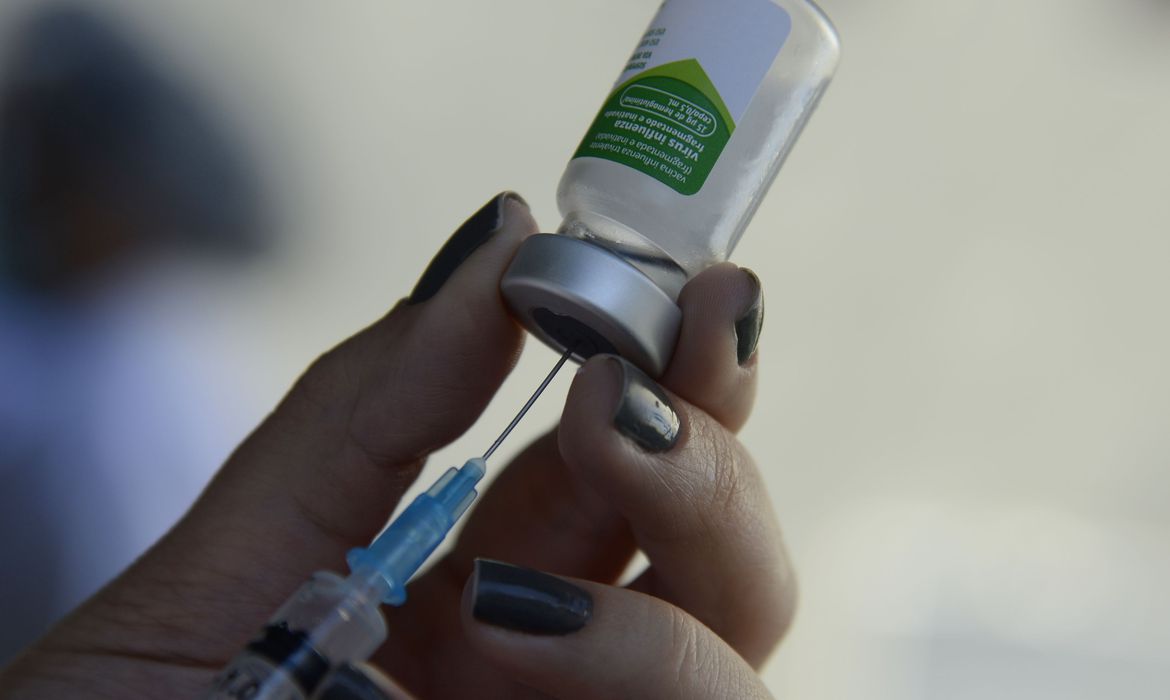 Mão retirando o líquido do frasco que armazena vacina contra gripe do Intituto Butantan