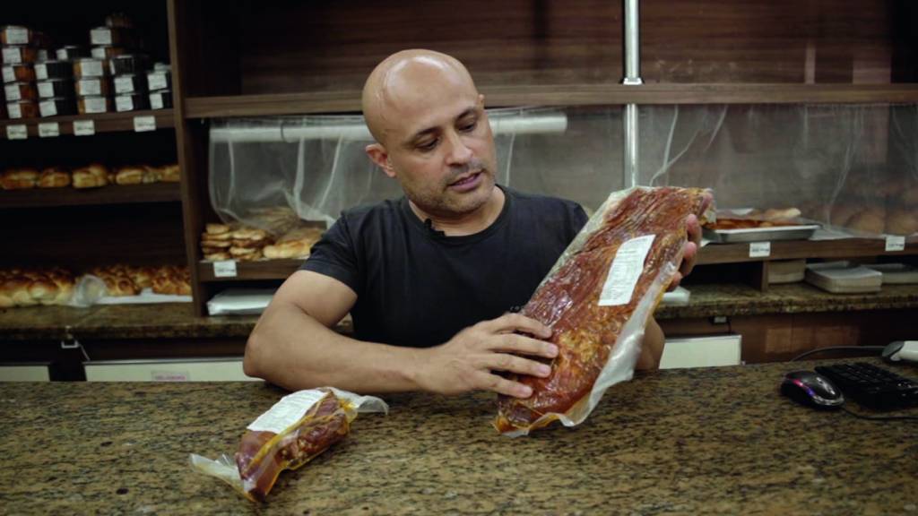 Homem branco, magro e careca sentado em uma mesa. Ele segura pacotes de carne de seu estabelecimento