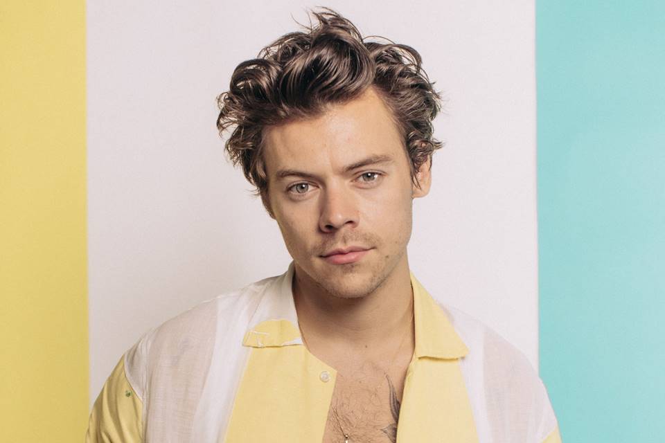 Harry Styles, de camisa colorida, posa em fundo com as mesmas cores de sua camisa: branco, amarelo e azul