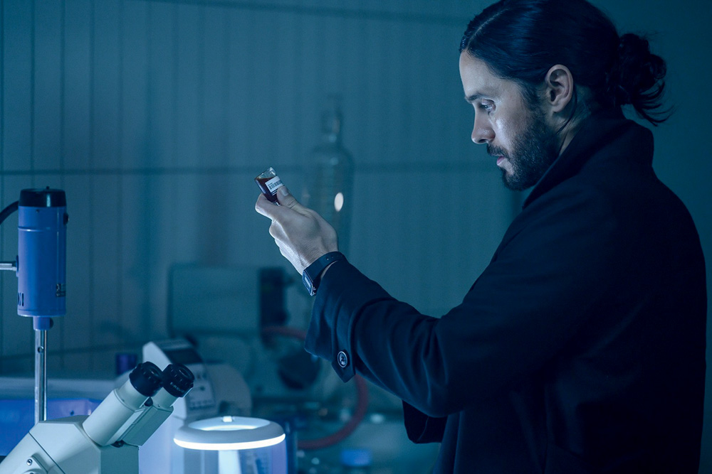 Imagem mostra homem de roupa preta e cabelo longo segurando um frasco em um laboratório