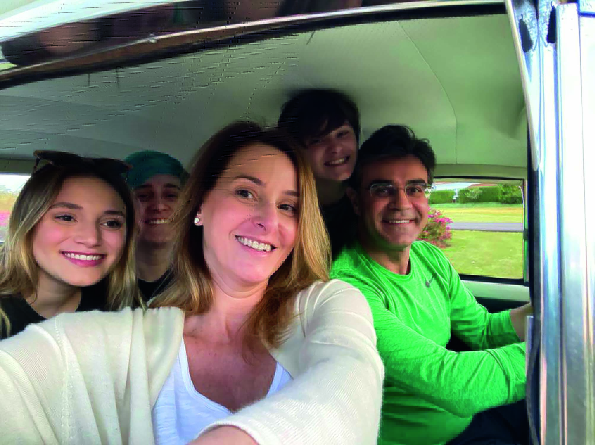 Imagem mostra família dentro de carro, todos sorrindo. Um homem de roupa verde está dirigindo, e uma mulher está tirando a foto. Os três filhos estão no banco de trás