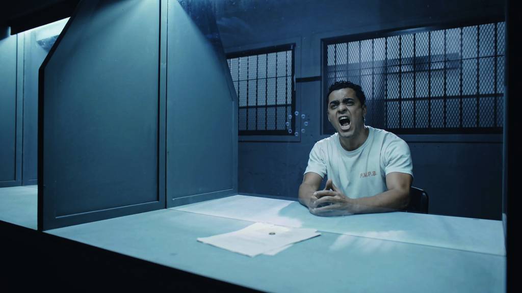 Imagem mostra homem gritando atrás de vidro. Ele está sentado e vestindo uma camiseta branca