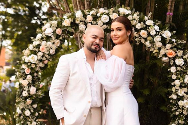 Imagem mostra o casal abraçado, vestidos de maneira formal, na frente de ambiente florido