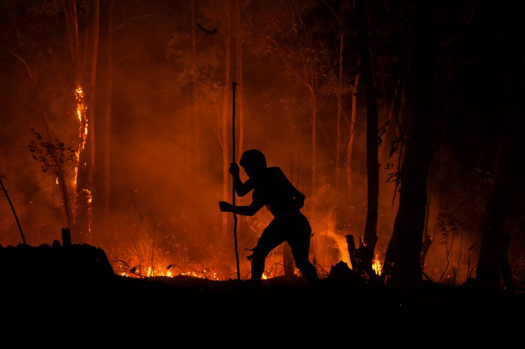 Imagem noturna mostra silhueta de pessoa com um incêndio ao fundo, em meio a uma floresta