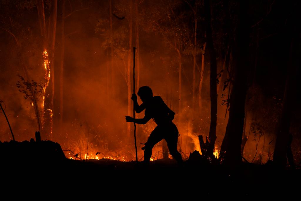 Fogo Imagem noturna mostra silhueta de pessoa com um incêndio ao fundo, em meio a uma floresta