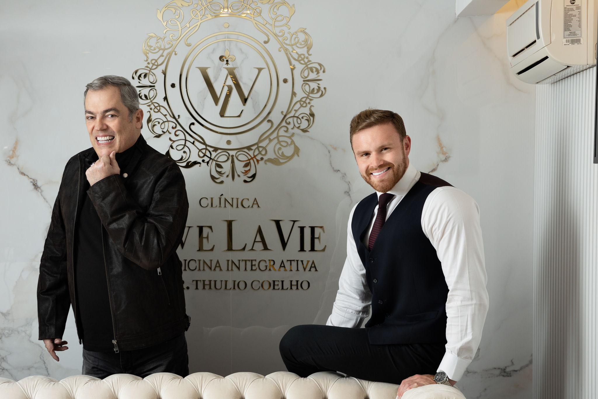 Marco Biaggi e Thulio Coelho posam lado a lado, ambos atrás de sofá rosa. Marco está de pé vestindo roupas pretas, Thulio está apoiado e veste camisa branca, gravata e colete preto.
