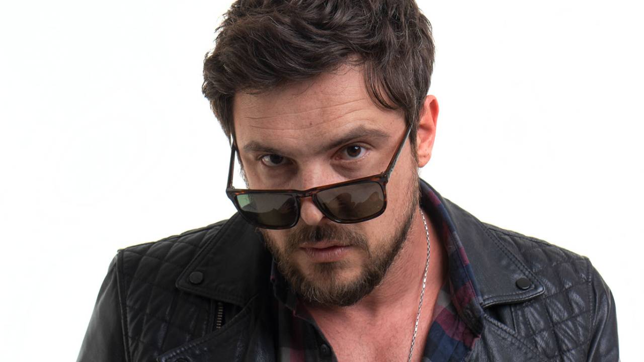 O ator e cantor Sérgio Guizé posa de jaqueta escura e óculos escuros apoiados no nariz.