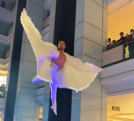 Frame de vídeo exibe bailarino com asas e calça legging branca suspenso no saguão do shopping
