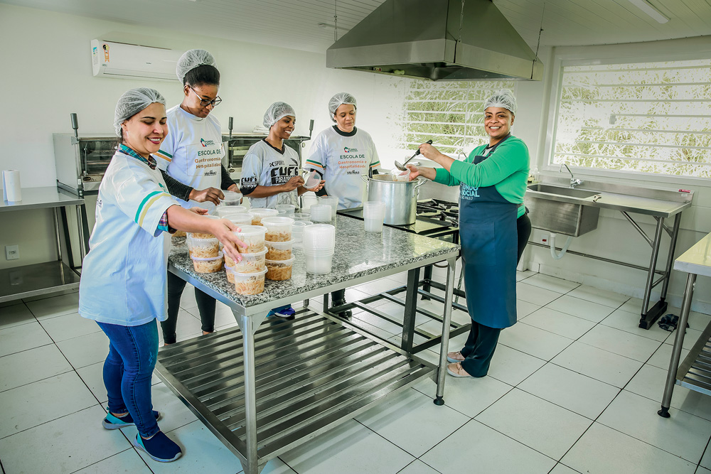 Quatro mulheres com toucas de cozinha estão em volta de uma mesa de metal em uma cozinha industrial