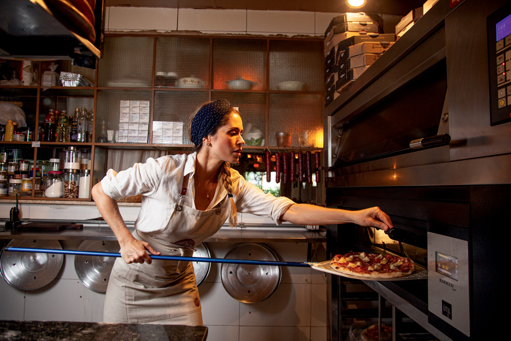 Imagem mostra mulher de camisa branca e avental bege colocando pizza em forno.