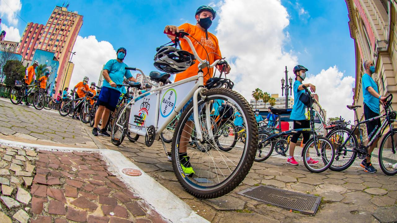 Imagem mostra diversas pessoas levando suas bicicletas, andando. Elas vestem roupas coloridas.