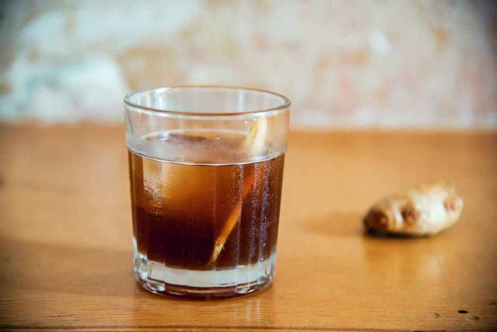 Drinque caraíva – Colchetes Copo de vidro com drinque marrom em uma mesa de madeira. Ao fundo, um peça de gengibre