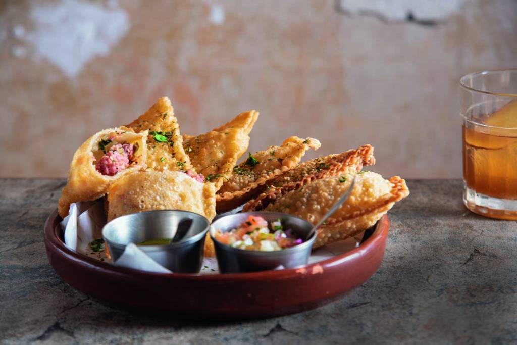 Pastéis de linguiça e de queijo – Colchetes Porção de pastéis de calabresa e queijo em um prato vinho. Dois potes de alumínio guardam molhos para acompanhar