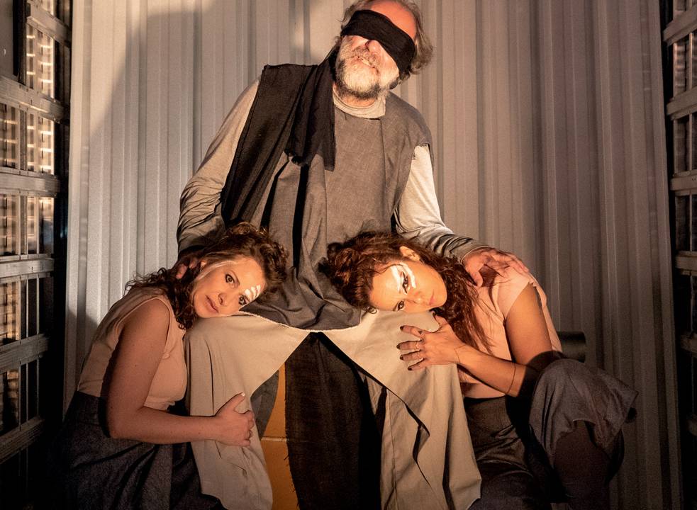 Um ator idoso com uma venda preta nos olhos está sentado enquanto duas atrizes deitam as cabeças em seu colo