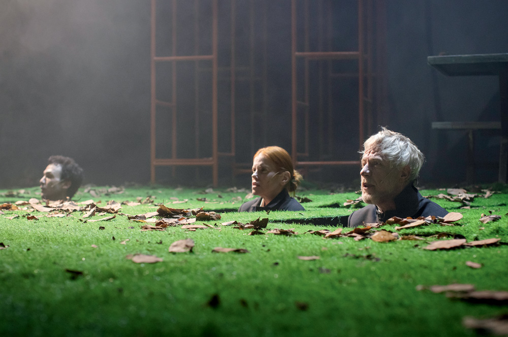 Três atores, dois homens e uma mulher, tem apenas as cabeças de fora em palco cheio de grama e folhas caídas