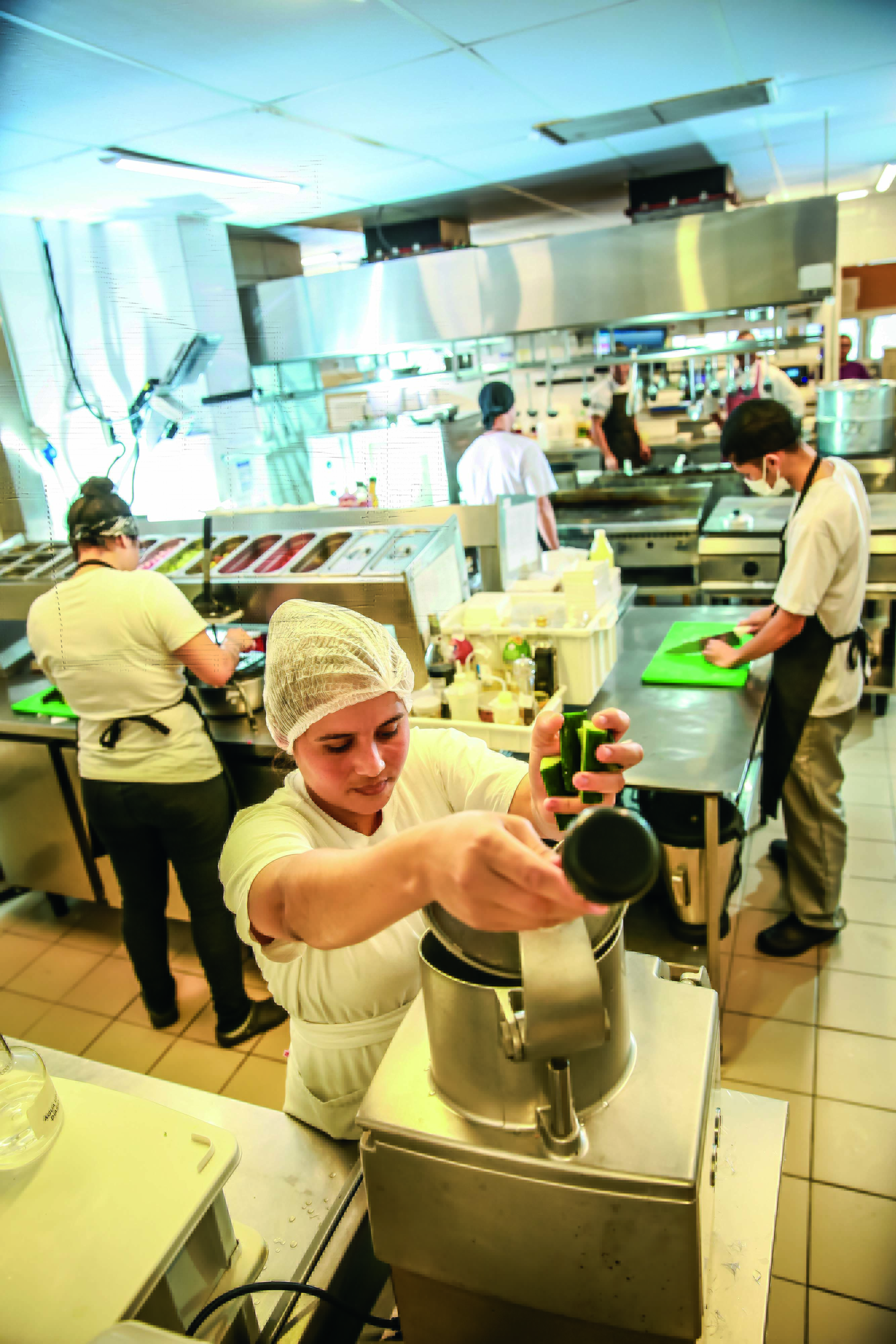 Imagem mostra cozinha com cozinheiros de roupas brancas e aventais. Em primeiro plano, uma mulher de touca colocando legumes em aparelho