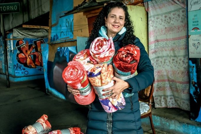 Érika é uma mulher magra e branca, de cabelos pretos e cacheados. Ela usa roupas escuras de frio e segura vários cobertores coloridos enrolados. Ela sorri para a foto e está em baixo de um viaduto