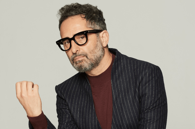 Imagem mostra homem de barba grisalha e óculos sentado, vestindo roupa de cor vinho com blazer preto, listrado.
