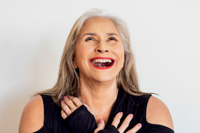 Imagem mostra mulher de cabelo branco sorrindo, olhando para cima, com as mãos sobre o peito