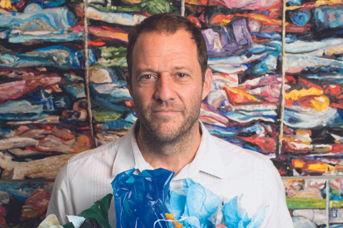 Imagem mostra homem segurando pedaços de plástico coloridos. Uma pintura colorida à óleo está pendurada ao fundo