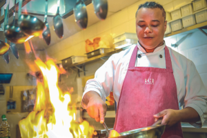 Imagem mostra homem cozinhando com frigideira na mão. Uma chama alta de um fogão é vista em primeiro plano