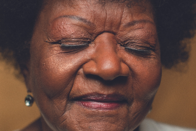Imagem mostra rosto de mulher idosa, com leve sorriso, de olhos fechados.