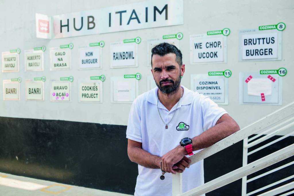 Imagem mostra homem de camiseta branca apoiado sobre corrimão. Ao fundo, parede com placas com nomes de restaurantes.