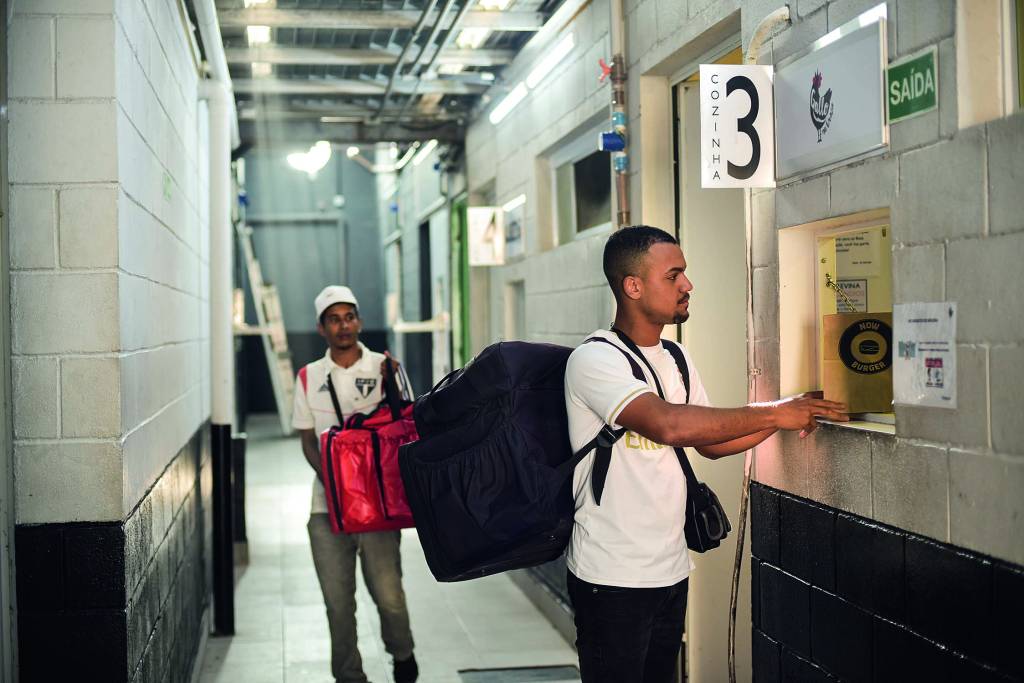 Imagem mostra dois homens em corredor, com bolsas de aplicativos de delivery