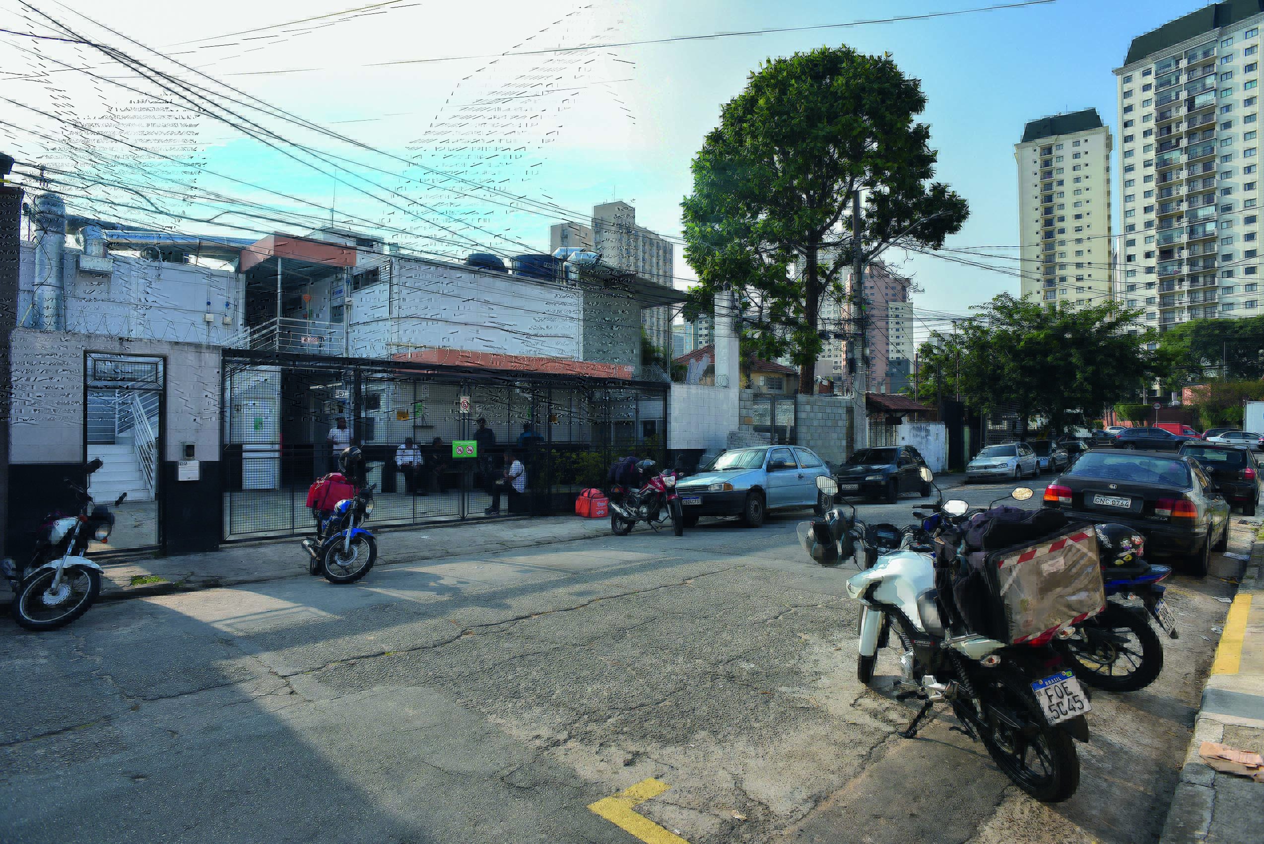 Imagem mostra rua com carros e motos estacionados. Ao fundo, um portão.