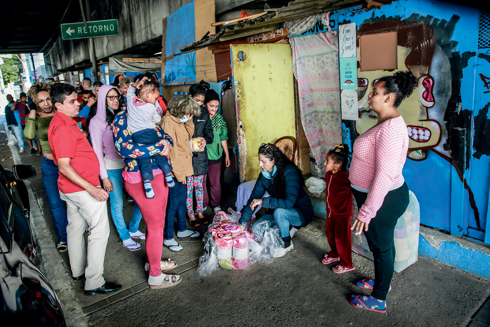 Erika, uma mulher branca e de cabelos escuros e cacheados, está agachada embaixo de um viaduto e distribui cobertores a pessoas em situação de rua em volta