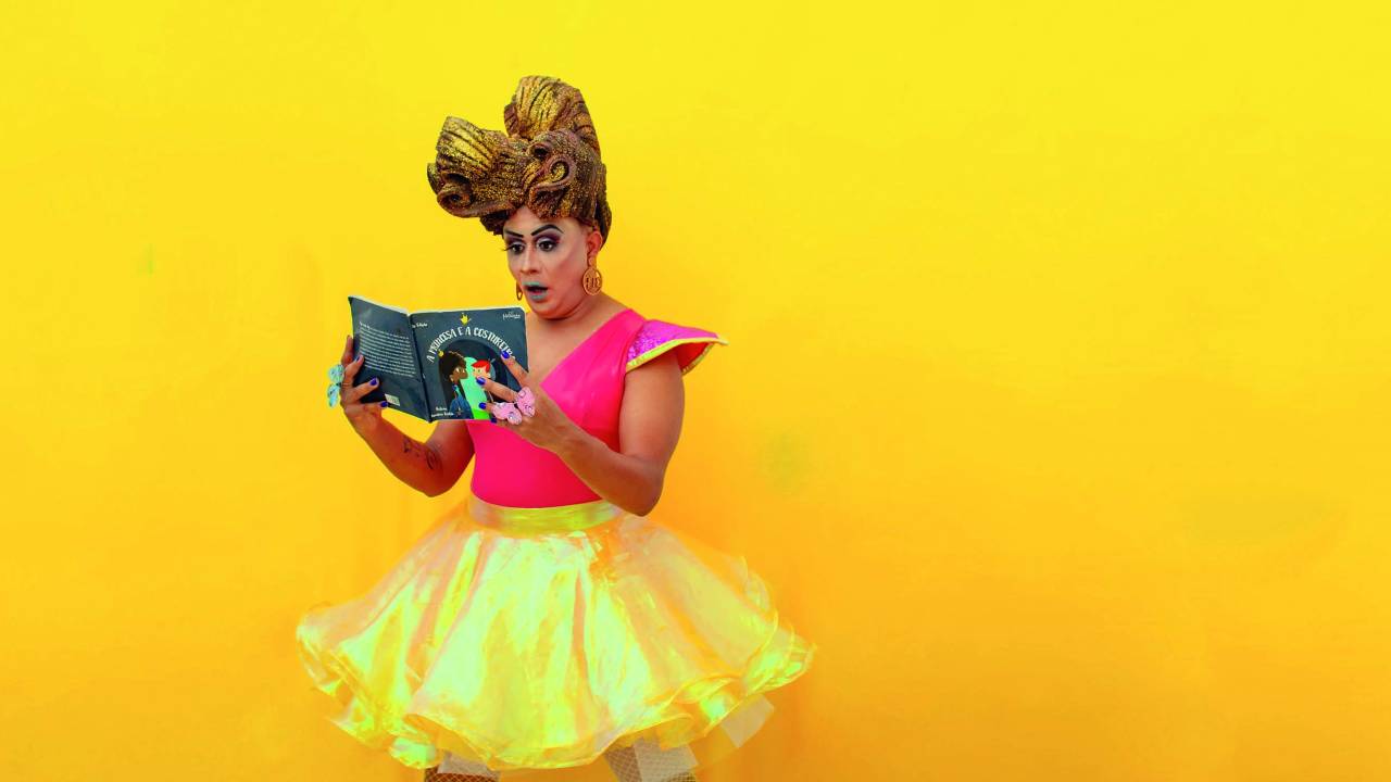 Helena Black, uma drag queen negra, usa uma roupa rosa de saia amarela e segura um livro infantil nas mãos