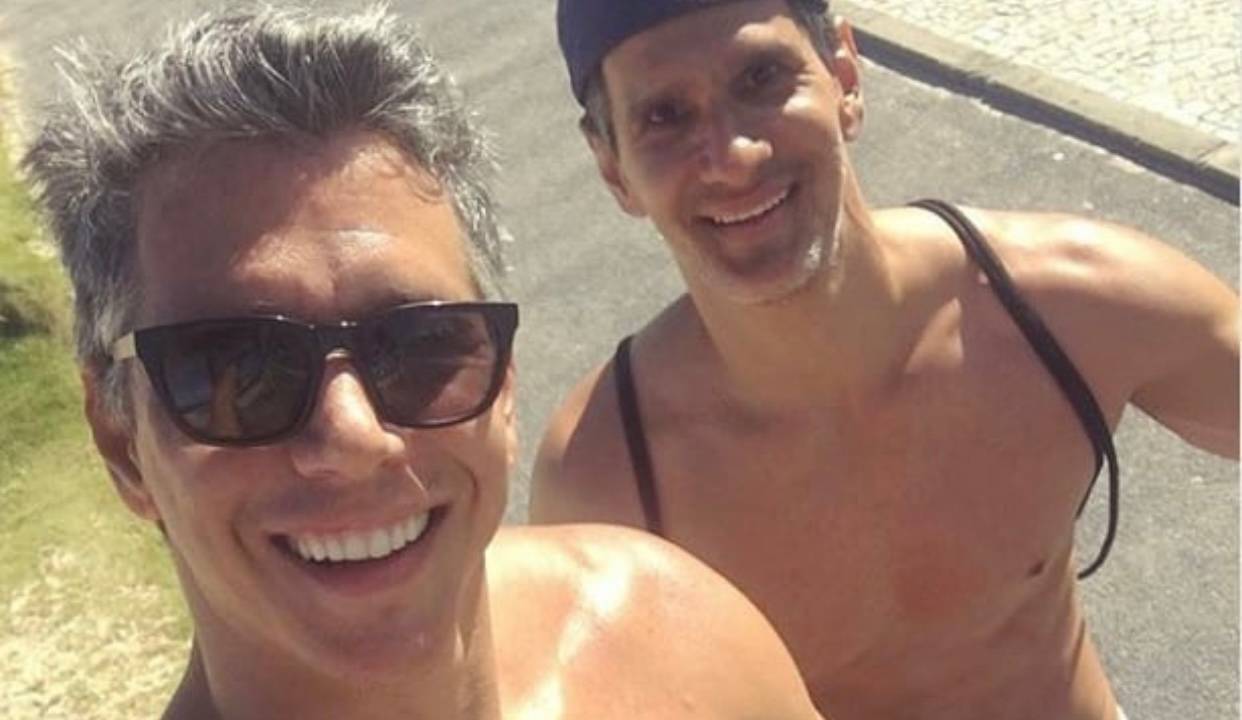 Imagem mostra dois homens, sem camisa, andando em calçada. Eles sorriem.