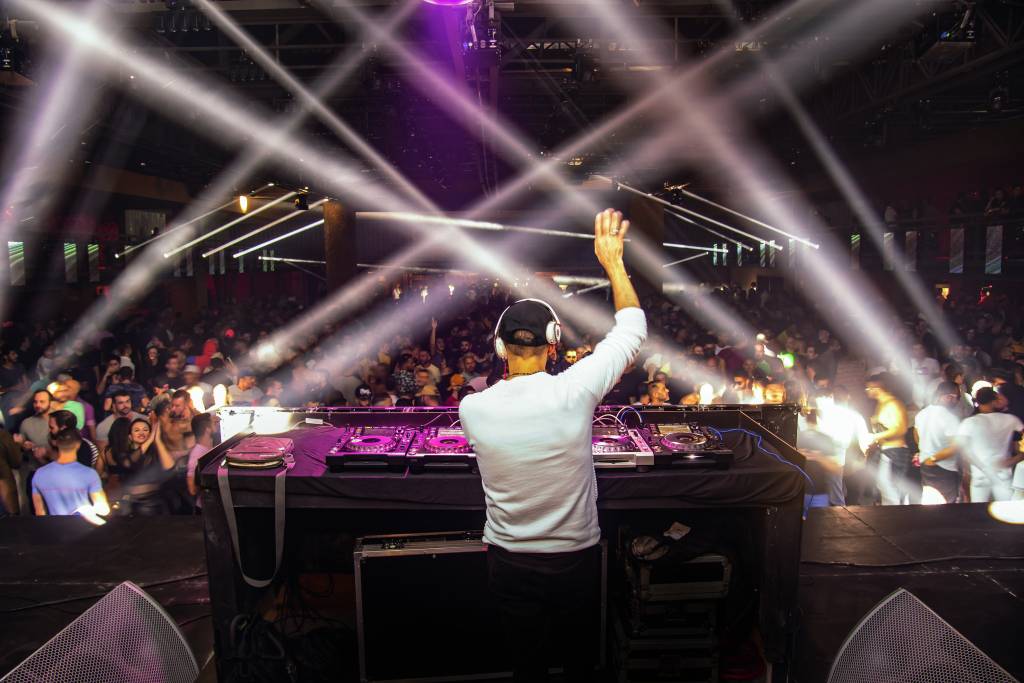 Imagem mostra Dj em mesa de som em cima de palco com multidão ao fundo. Luzes brancas e roxas iluminam o ambiente