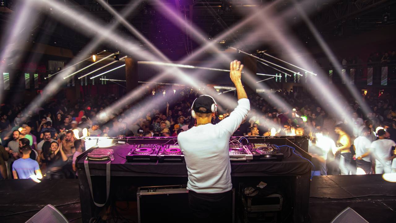 Imagem mostra Dj em mesa de som em cima de palco com multidão ao fundo. Luzes brancas e roxas iluminam o ambiente