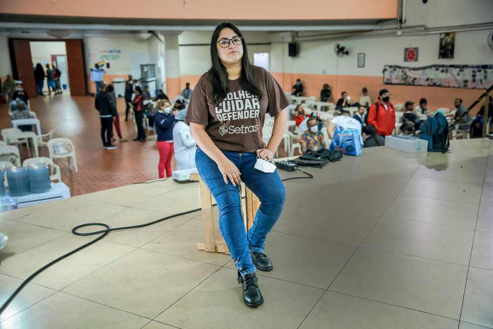 Léa é uma mulher branca, magra, de cabelos lisos e escuros. Usa óculos. Está sentada em um banco no prédio do Chá do Padre