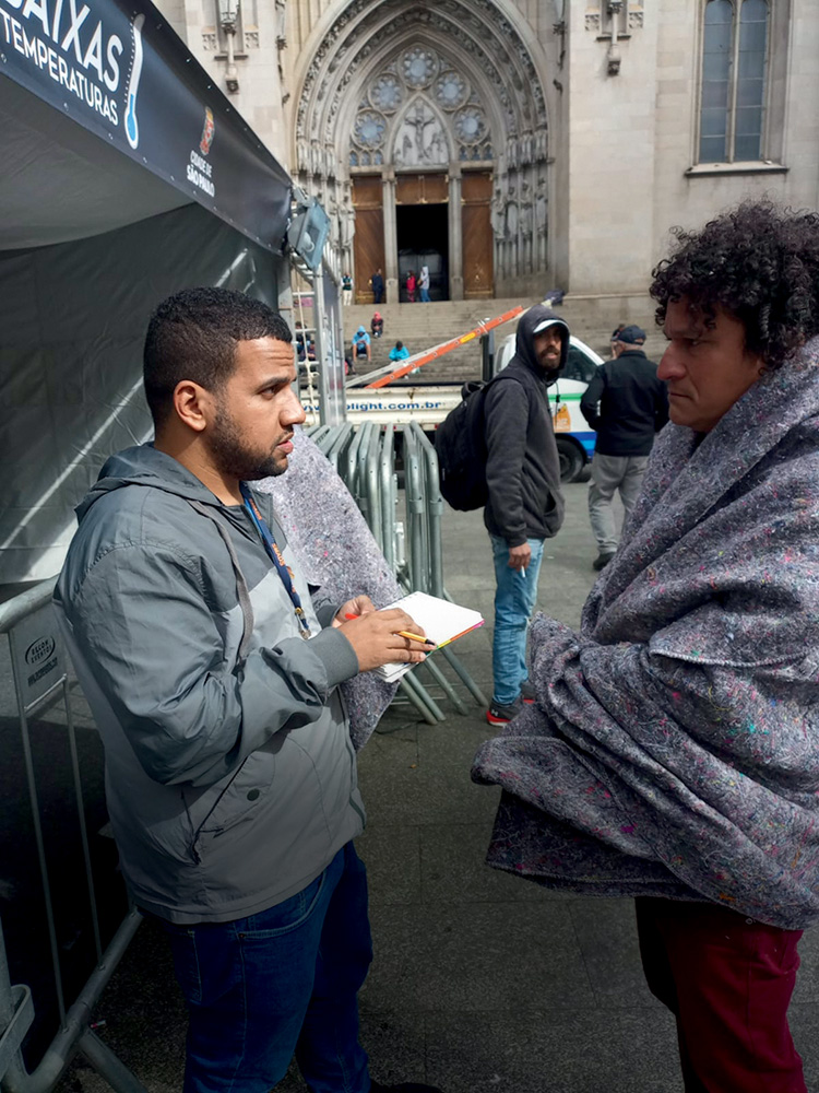 Um homem pardo, de barba e cabelos escuros, conversa com um homem também pardo em situação de rua. Eles estão na Praça da Sé e o morador de rua está envolto em um cobertor