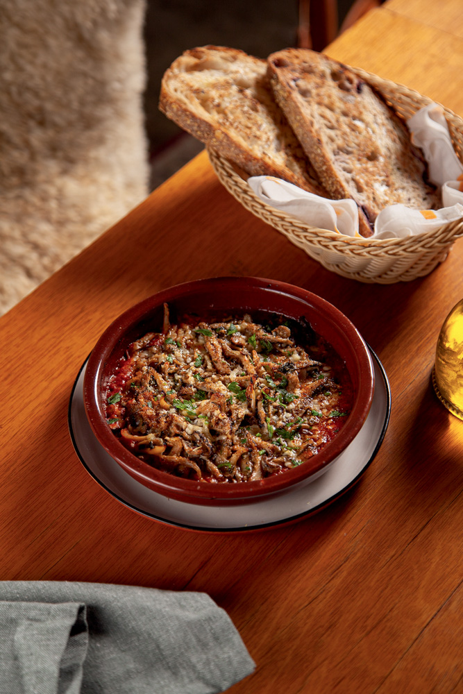 Imagem mostra prato com cogumelos sobre mesa de madeira. Na parte superior da imagem, é possível ver uma cesta de pães.