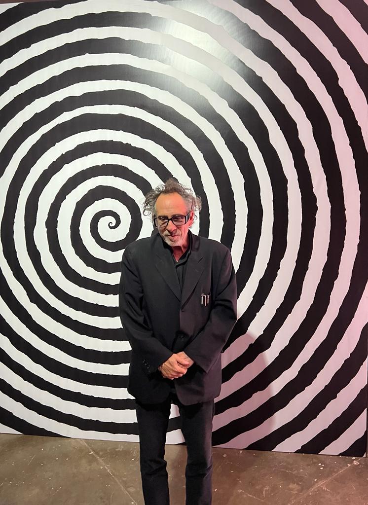 Mostra Tim Burton (VEJASP) (1) O cineasta Tim Burton posa de terno preto em frente a fundo com espécie de caleidoscópio preto e branco.