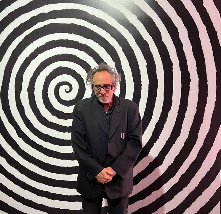 O cineasta Tim Burton posa de terno preto em frente a fundo com espécie de caleidoscópio preto e branco.