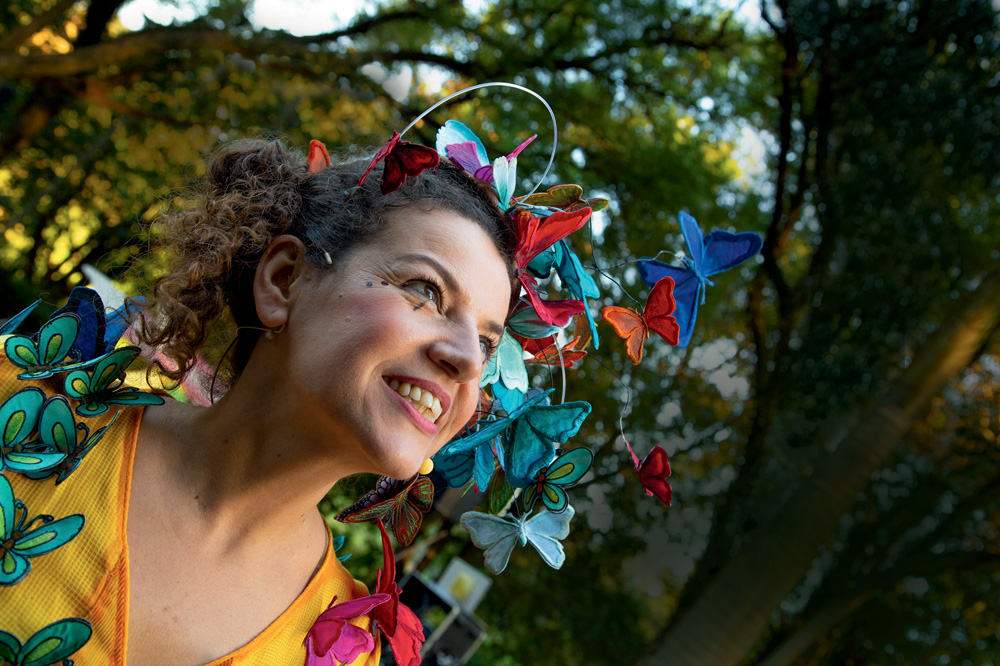 Eloisa é uma mulher branca e de cabelos castanhos cacheados. Ela aparece do peito para cima, sorrindo, e veste um vestido e uma tiara cobertos por borboletas coloridas de pano