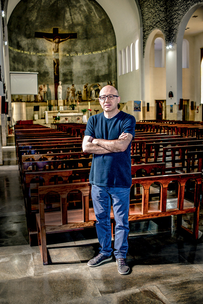 Um padre branco, magro e careca posa em frente aos bancos de uma Igreja Católica. Ele usa camiseta azul marinho e calça jeans